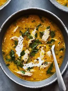 A steaming bowl of chicken soup beside orange citrus halves and a small bowl of yogurt on a kitchen table
