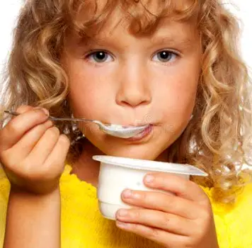 A young child at a kitchen table eating a spoonful of yogurt with an ice pop, cup of diluted juice, and applesauce nearby
