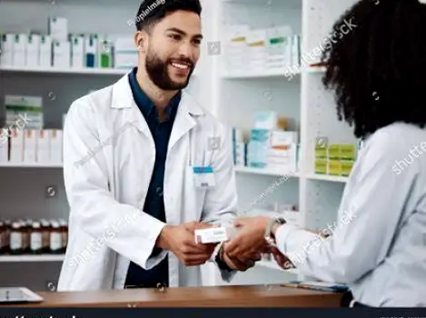 A pharmacist and a customer standing across a pharmacy counter, with the customer holding a medicine box while the pharmacist points toward product information