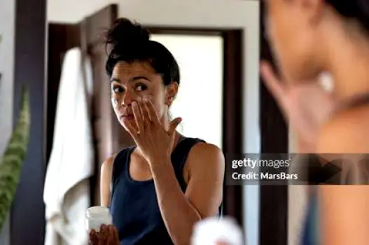 Woman with dry skin on her cheeks applying a small amount of fragrance-free face cream in front of a bathroom mirror